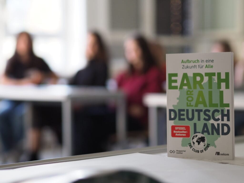 A copy of Earth for All Germany book against a blurred background of young participants in a workshop