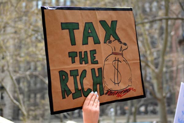 A hand holding a cardboard handwritten sign saying Tax the Rich