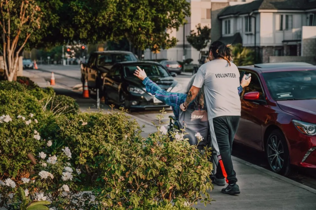 A man with a 'volunteer' tshirt pushing a person in a wheelchair past a garden