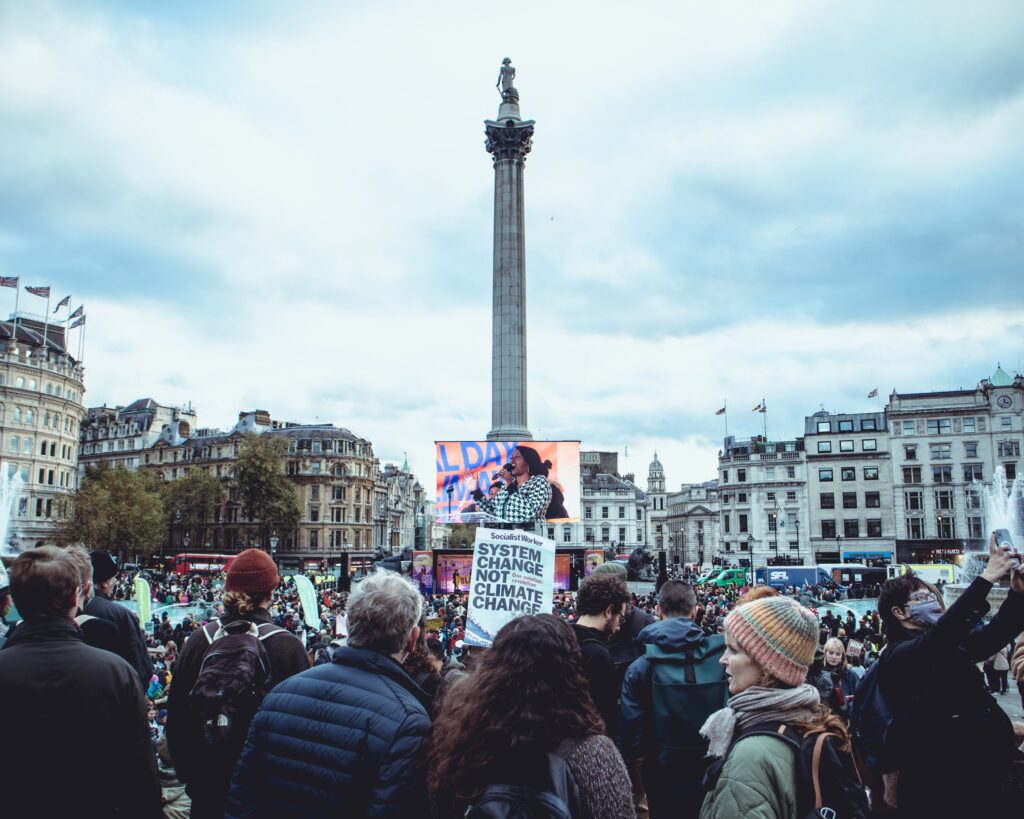 A crowd gathers in Trafalgar Square, London, facing a large screen showing a speaker, with a protest sign reading System Change Not Climate Change.