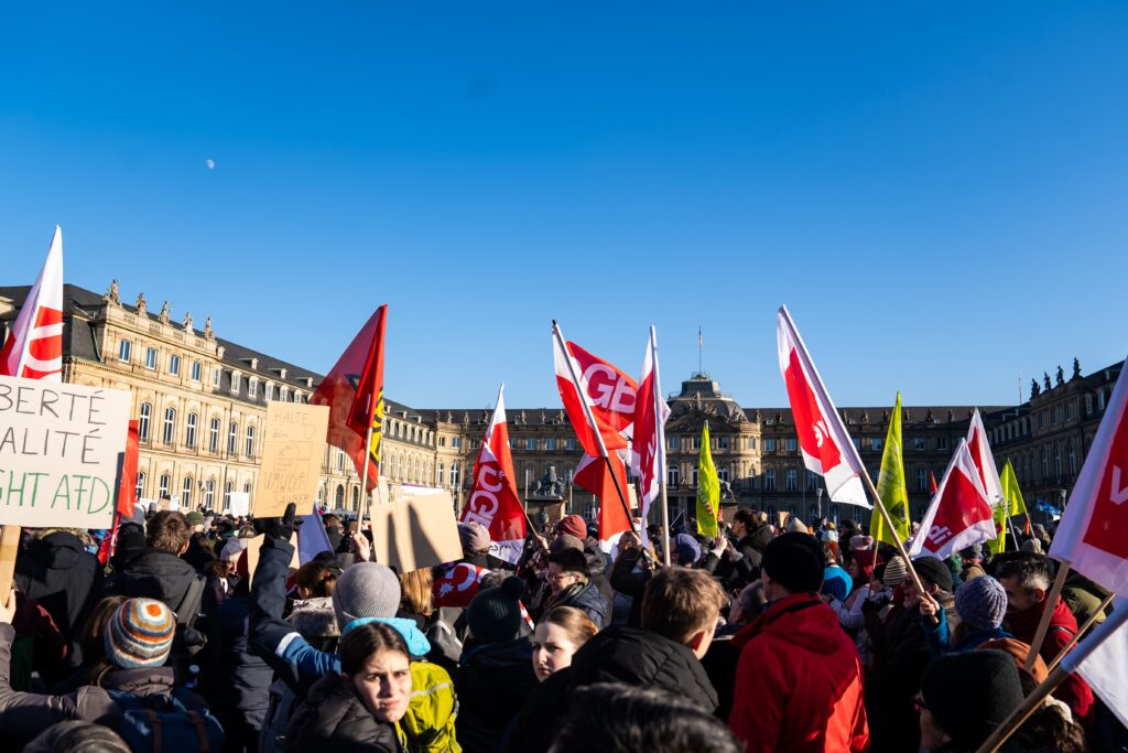 A large crowd gathers in a square, holding various flags and signs, with historic buildings in the background under a clear blue sky.
